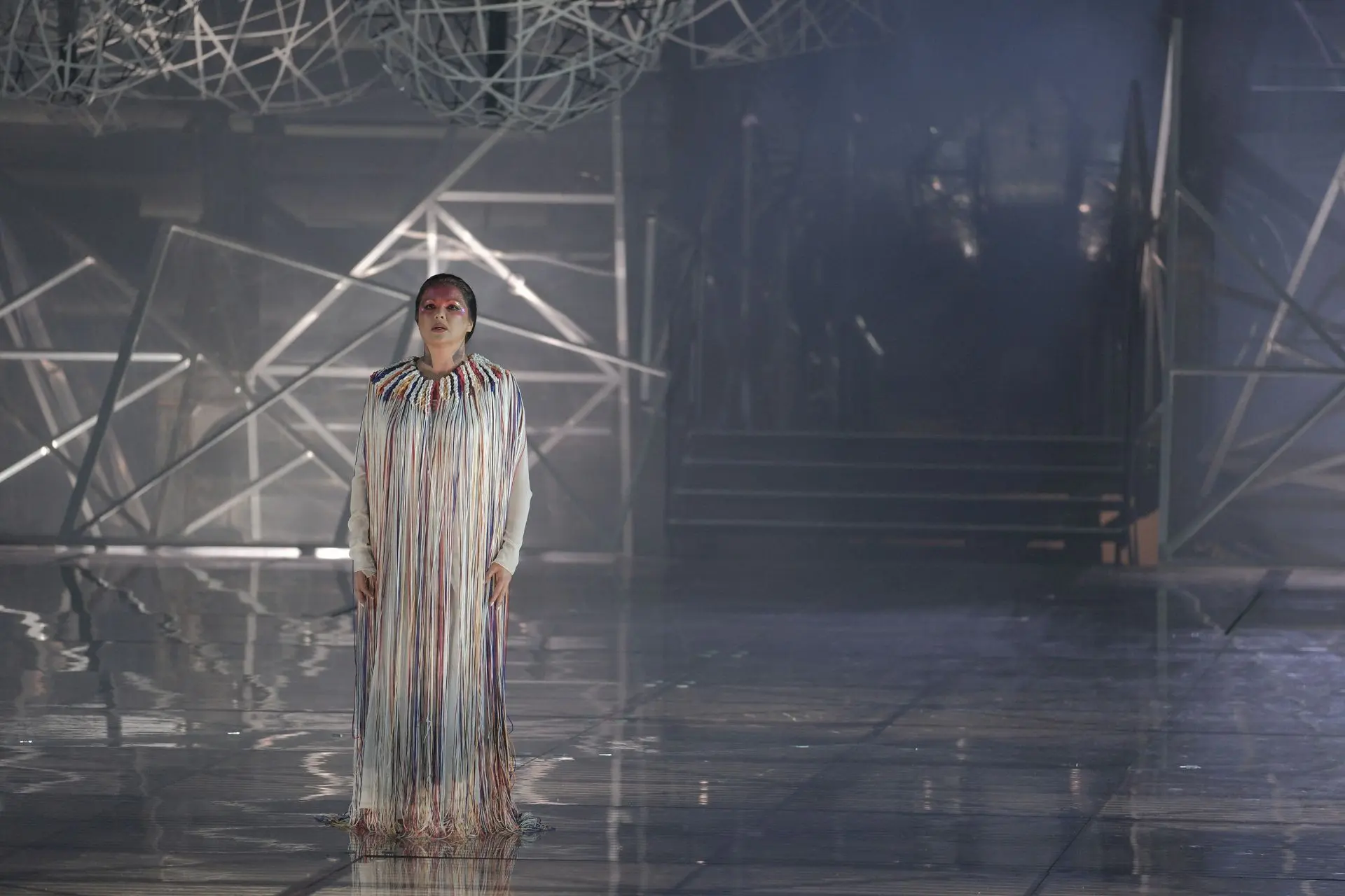 She is standing at the center of the stage. She is wearing a long sleeve white suit completely covered by a sleeveless robe. The robe has a wide collar with white, red, blue and yellow stripes. Strings of the same colors descend from the collar covering the figure of Aida to her feet. She has black hair with a few red locks. It is pulled back and held in place by a chignon on the nape of her neck. On her forehead, she is wearing light red and purple makeup covered with multicolored glitter. The sides of her neck are decorated with silver glitter that forms a sort of upside down “V”. 