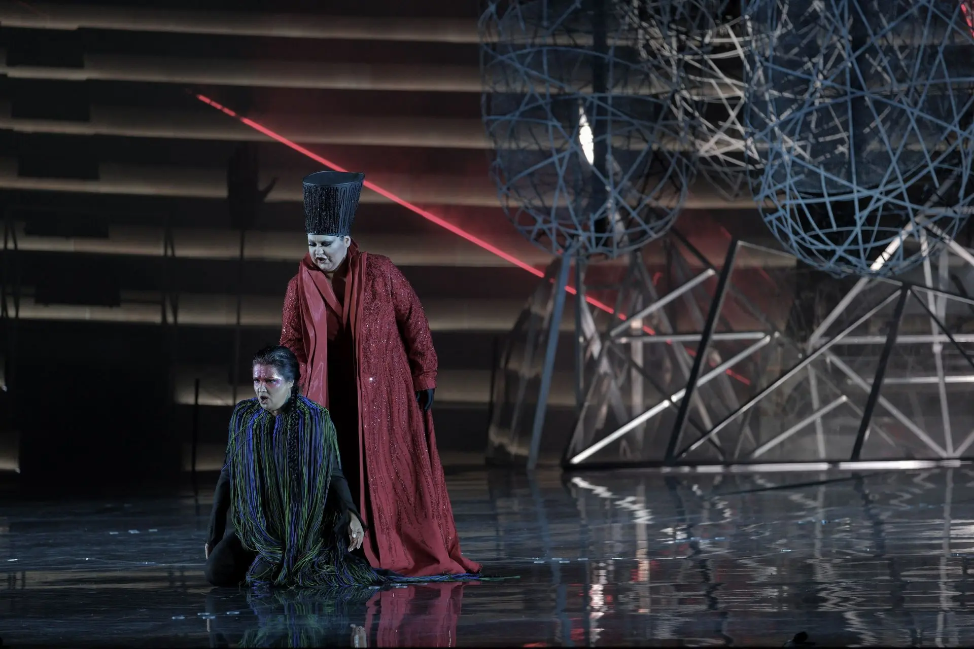 Aida and Amneris, Act II. Aida is on her knees at the center of the stage. Behind her stands Amneris. Aida is wearing a long sleeve black suit, completely covered by a sleeveless robe. The robe has a wide collar with light green, dark green and blue strips. Strings of the same colors fall from the collar covering the figure of Aida to her feet. She has black hair tied in a long braid. She has makeup on her forehead and the sides of her neck are decorated with glitter. Amneris is wearing a floor length Bordeaux robe. It is open in the front and accented with strings of stone of the same color that reflect the light. Underneath she is wearing a floor length Bordeaux suit. On her head, she is wearing a tall, cylindrical, black headdress that widens slightly at the top. It is decorated with strings of black stones. She is also wearing black gloves and black makeup on her eyelids.