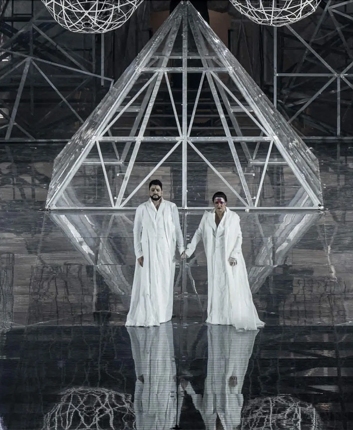Aida and Radamès, Act IV. They are standing at the center of the stage in front of the pyramid and are holding hands. They are wearing loose fitting white robes that are floor length and open in the front. Underneath, both are wearing floor length garments made of the same color. Aida has her hair pulled back and held in place by a chignon on the nape of her neck. On her forehead, she is wearing light red and purple makeup covered with multicolored glitter. Radamès has short black hair and a beard. At the center of his forehead is a small black line.