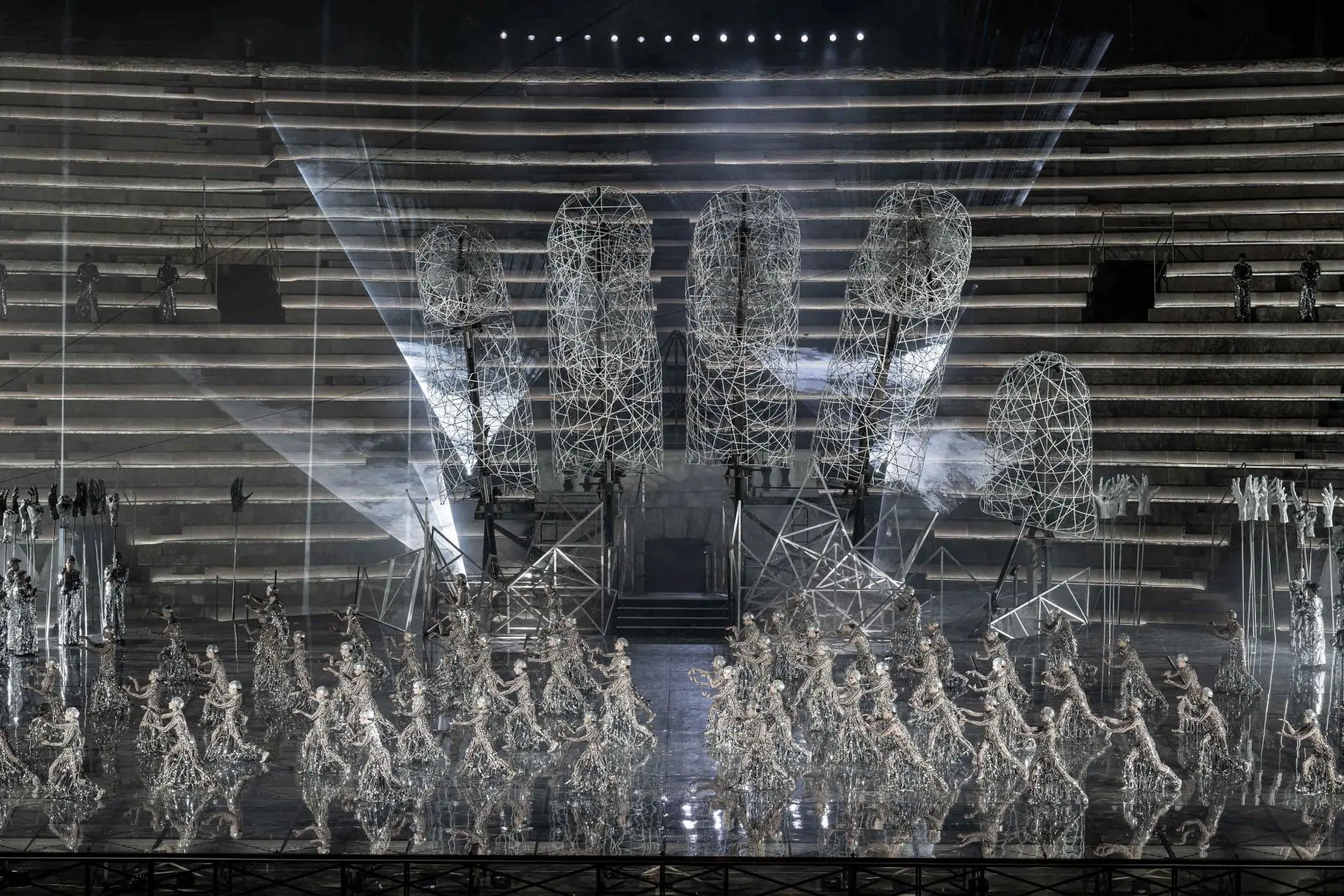 The stage is again dominated by the large hand, illuminated by beams of white light and surrounded by dense clouds of smoke. At the back of the stage, the spears with white hands can be seen to the right while the spears with the black hands are to the left. They are all made of papier-mâché. In front of the large hand, male and female dancers occupy the entire stage. They are standing and facing the right with both their right leg and their arms extending forwards. They are wearing tight beige colored suits decorated with designs of black hieroglyphs. On top of their suits, they are wearing sparkling silver netting accented with crystals with silver caps on their heads. 