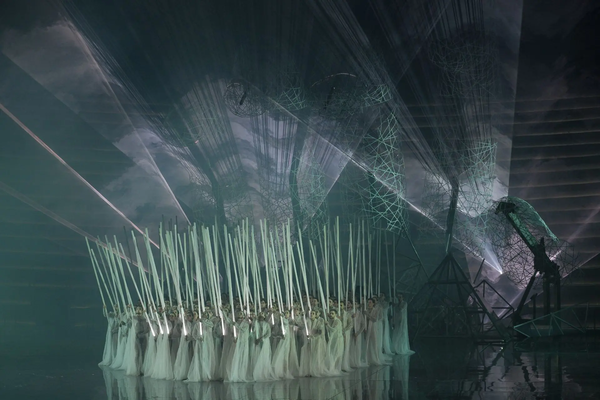 The stage is again dominated by the large hand, illuminated by beams of white light and surrounded by dense clouds of smoke. The male and female dancers are standing next to each other and occupy the center of the stage. On top of their suits they are now wearing long white robes made of tulle. In their hands, they are holding long white illuminated staffs, similar to laser swords. They are held pointing upwards. 
