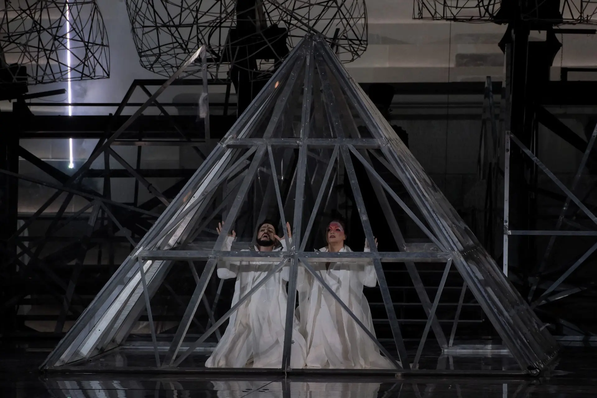Radamès and Aida are on their knees side by side within an iron structure in the form of a pyramid that recalls the one outside the Louvre. Both are dressed in white and hold their hands up in front of them. Aida is wearing light red and purple makeup on her forehead covered with multicolored glitter, while Radamès, has a small black line at the center of his.