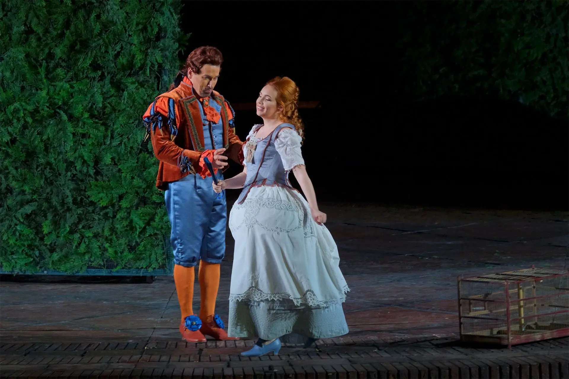 Figaro and Rosina are standing next to each other at center stage in front of the labyrinth. Figaro is wearing a bright blue satin suit consisting of a buttoned waistcoat and knee length breeches with orange stockings below them. Over the waistcoat he is wearing an orange velvet jacket that is open in the front. The jacket is shorter and straight in the front and longer and pleated in the back. The shoulders are padded and accented with blue and orange inserts, while the wrists and collar have bright orange ruffles. On his feet he is wearing squarish shoes with a low heel and a large blue flower at the neck of the foot. He is wearing a chestnut colored wig with the hair pulled back and tied in a tail with a ribbon. Rosina is wearing a gown made of light cotton with a wide skirt that falls to her ankles. The top consists of reinforced bodice that is a darker shade of sky blue with red trim. The sleeves are short and light blue. The skirt is light blue, wide and layered. The top layer is accented with lace. On her feet she is wearing squarish shoes with a low heel. Her is a light chestnut brown color, almost orange. It is pulled back on the nape of her neck and falls down to her shoulders. She is holding a blue hand fan that is closed. 