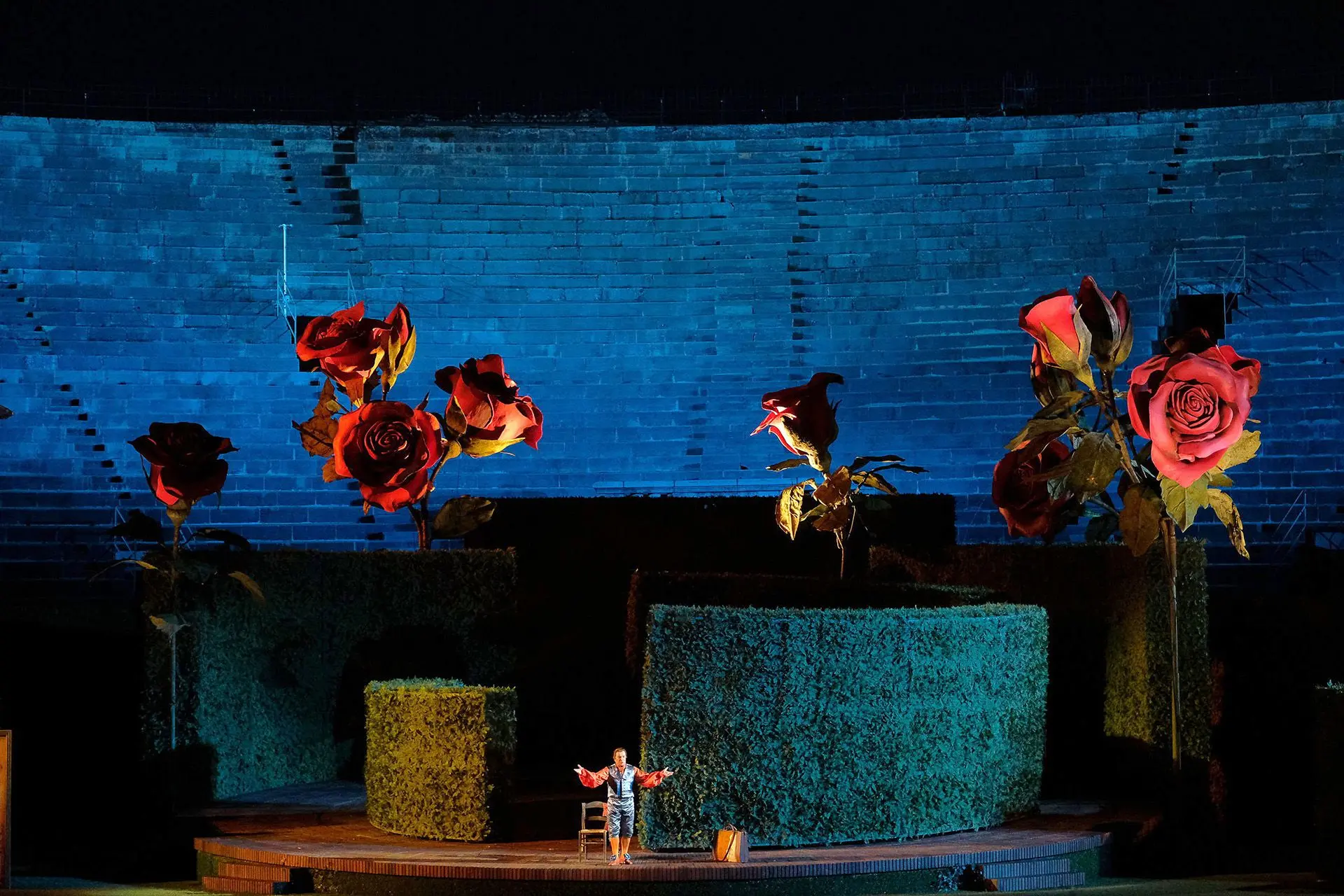 The stage is dominated by a part of a circular labyrinth of concentric semicircles that vary in height. The semicircles are made up of enormous green hedges set upon a raised circular base. They are surrounded by 8 giant red roses made out of papier-mâché. The roses have long green stems coming up from the floor of the stage with leaves of the same color. Figaro is in front of the labyrinth at center stage. Behind him is a wooden chair with a straw seat. To his left is a piece luggage on the ground. Given the colossal dimensions of the labyrinth and the roses, Figaro appears to be minute in comparison.
