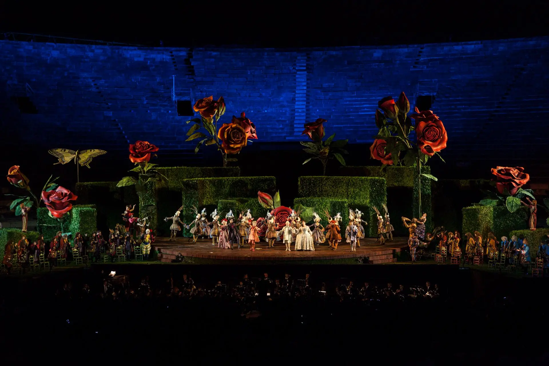 The stage is entirely taken up by the labyrinth resting on the circular base. It is surrounded by 12 roses, and, on the left, there is a giant colorful butterfly made out of papier-mâché. Its wings are open and it is positioned on top of a long pole fixed to the floor of the stage. In front of it stand approximately 20 danseurs and ballerinas dressed in splendid eighteenth-century costumes, along with the rest of the characters in the opera who are also wearing clothing of the same period. Among them are also the Count and Rosina who are holding each other’s hands. At the far left and right of the scene, there are approximately 30 chairs on each side. Some are bright green in color and others are orange. Near the chairs stands the chorus.