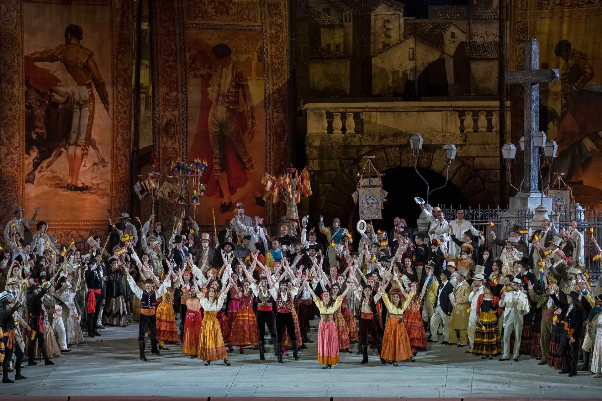 La scena rappresenta la piazza antistante la Plaza de Toros nel giorno della corrida. Al centro si trovano dei danzatori e delle danzatrici di flamenco, circondati da una folla composta da donne e uomini, molti dei quali sventolano delle bandierine della Spagna, gialle e rosse. Tra loro, anche dei venditori ambulanti; questi ultimi sorreggono degli espositori con le bandierine e con delle girandole colorate. Sulla destra della piazza si trova una croce votiva posizionata su una base di pietra, circondata da un’inferriata e da alcuni lampioni. Sul fondo del palcoscenico, dietro alla folla, l’ingresso della Plaza de Toros è costituito da un arco in pietra sormontato da una balaustra. L’ingresso è circondato da grandi locandine di corride, sempre in stile liberty.