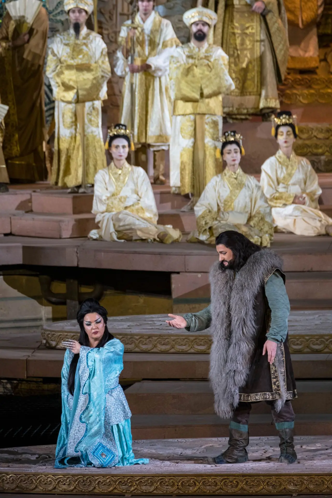 At center stage kneels princess Turandot. To her left is Calàf, dressed the same as in the previous picture. Turandot is wearing a pale ice blue robe that falls to her feet, with sleeves that widen at her wrists. They are shiny and accented by rolling embroidery in sky blue. The shoulders are padded, resembling armor. The front of the robe is decorated with shiny sky-blue bands. At her feet, only the toes of her shoes are visible, and they are same color as the robe. On her head she is wearing a headdress in the shape of silver sunburst. The tips of the rays are blue.
