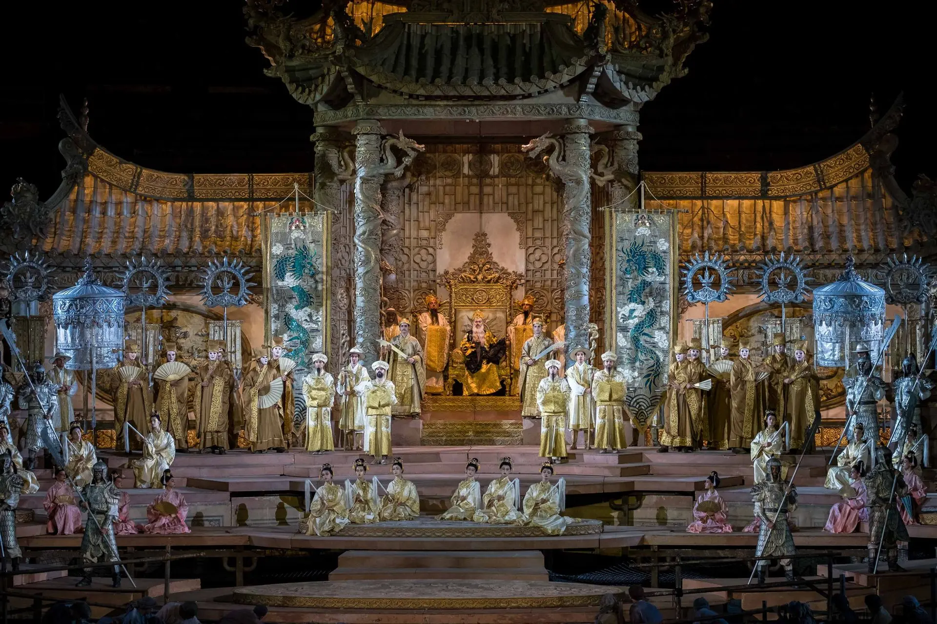 The stage is dominated by the palace of emperor Altoum. The gold and silver of the building glitter with jade green reflections. The main part of the building is formed by the baldacchino above the throne. It recalls the Baldacchino of Saint Peter’s Basilica by Bernini. It is supported by four twisted Solomonic columns with dragons coiled around them. Sitting on the throne is emperor Altoum. To the right and left of the baldacchino stand the banners of the emperor, one on each side. They are silver and gold with a blue dragon emblazoned on them. In front there are steps where the crowd stands. There are ballerinas in pink with parasols, supernumeraries in white with plumes, dignitaries in beige with fans, and concubines dressed in clothes made of shiny cloth. This scene creates an effective contrast between the imperial space and that of the commoners, which appears even more shabby and listless.
