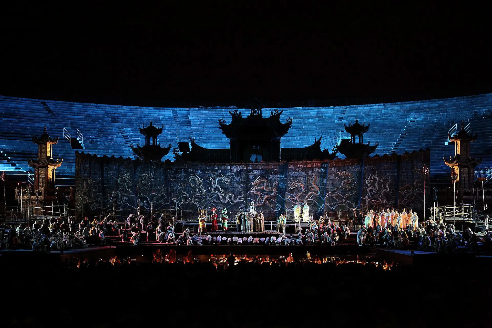 The stage is dominated by a structure made of wood where the people of Peking are gathered. Some are sitting, others crouching, and still others prostrate. On the central walkway stand the three ministers of state Ping, Pong e Pang, dressed in yellow, red and green, with a dominant color for each. Princess Turandot is there dressed in a silvery suit similar to armor. In the back, along the brick wall with the dragons, you can discern pagodas and towering buildings with overlapping roofs: a forbidden city hidden from view. In the background are the tiers of the arena.
